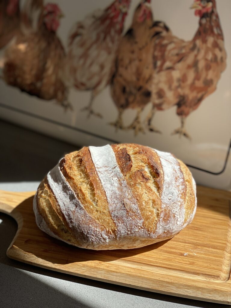 A sourdough loaf of bread sitting on a cutting board with a picture of chickens behind it.
